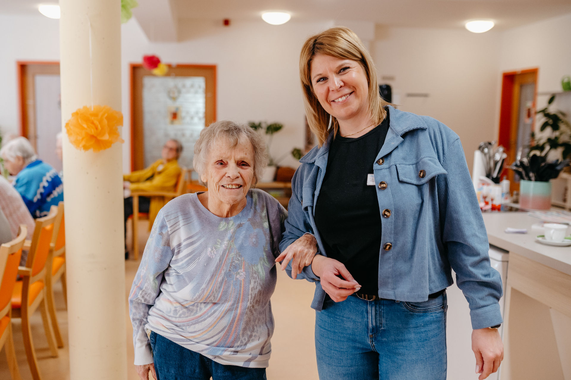 Ältere Frau steht neben einer jüngeren Mitarbeiterin und hält deren Arm, beide lächeln in einem hellen Gemeinschaftsraum der Einrichtung.