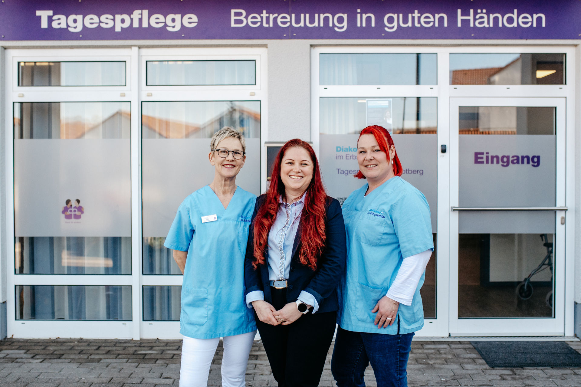 Drei lächelnde Mitarbeitende stehen vor dem Eingang der Tagespflege; zwei tragen hellblaue Pflegeuniformen, die mittlere Person Business-Kleidung.