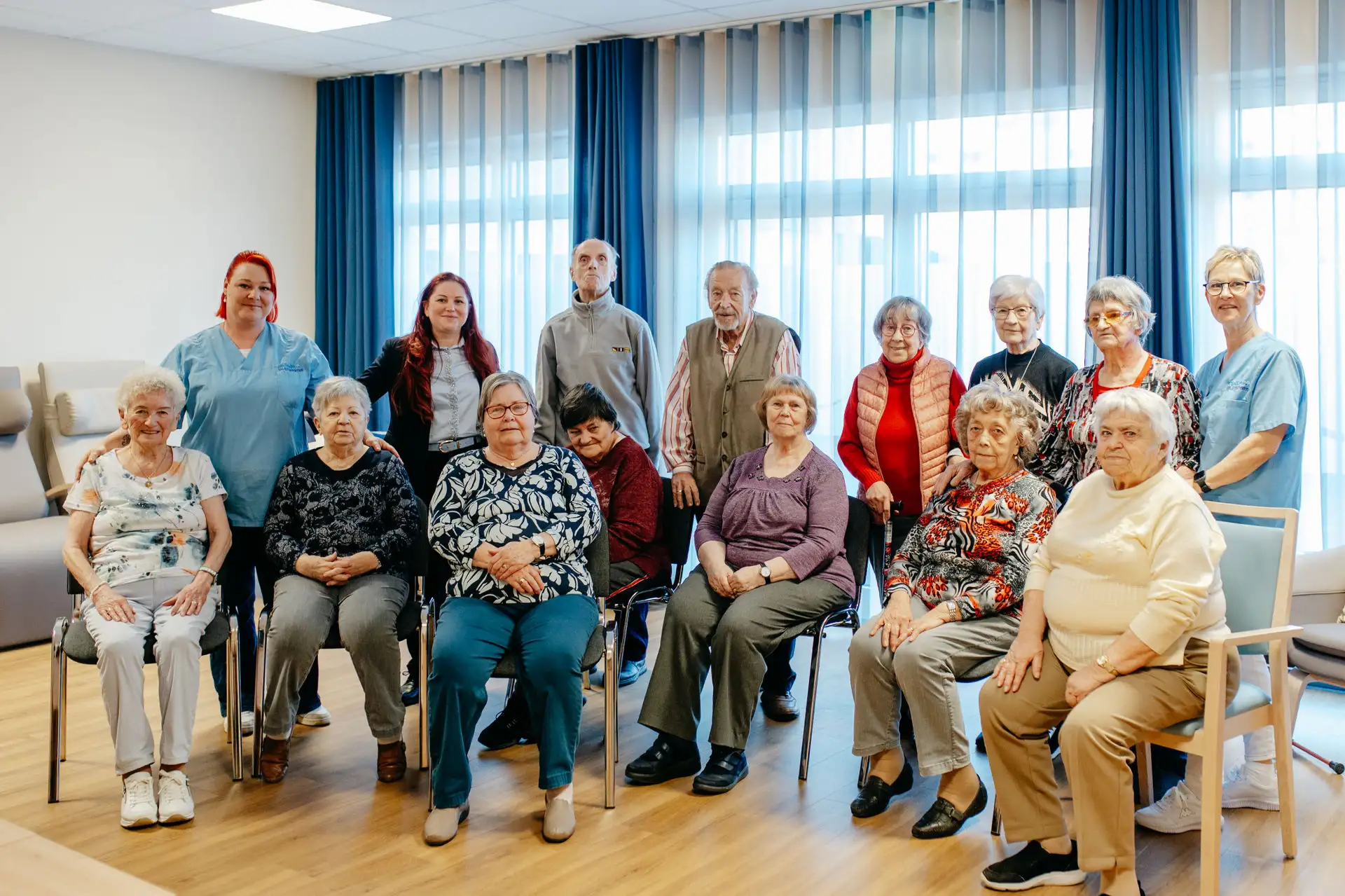 Ältere Frauen und Männer sitzen und stehen in einem Gruppenraum, begleitet von Pflegerinnen; die Stimmung wirkt ruhig und gemeinschaftlich.