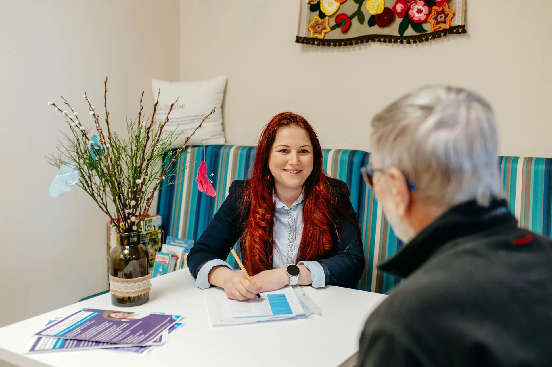 Beraterin mit roten Haaren sitzt lächelnd an einem Tisch und füllt mit Bleistift Unterlagen aus, während ein älterer Klient ihr gegenüber sitzt; helle, freundlich dekorierte Beratungsumgebung.