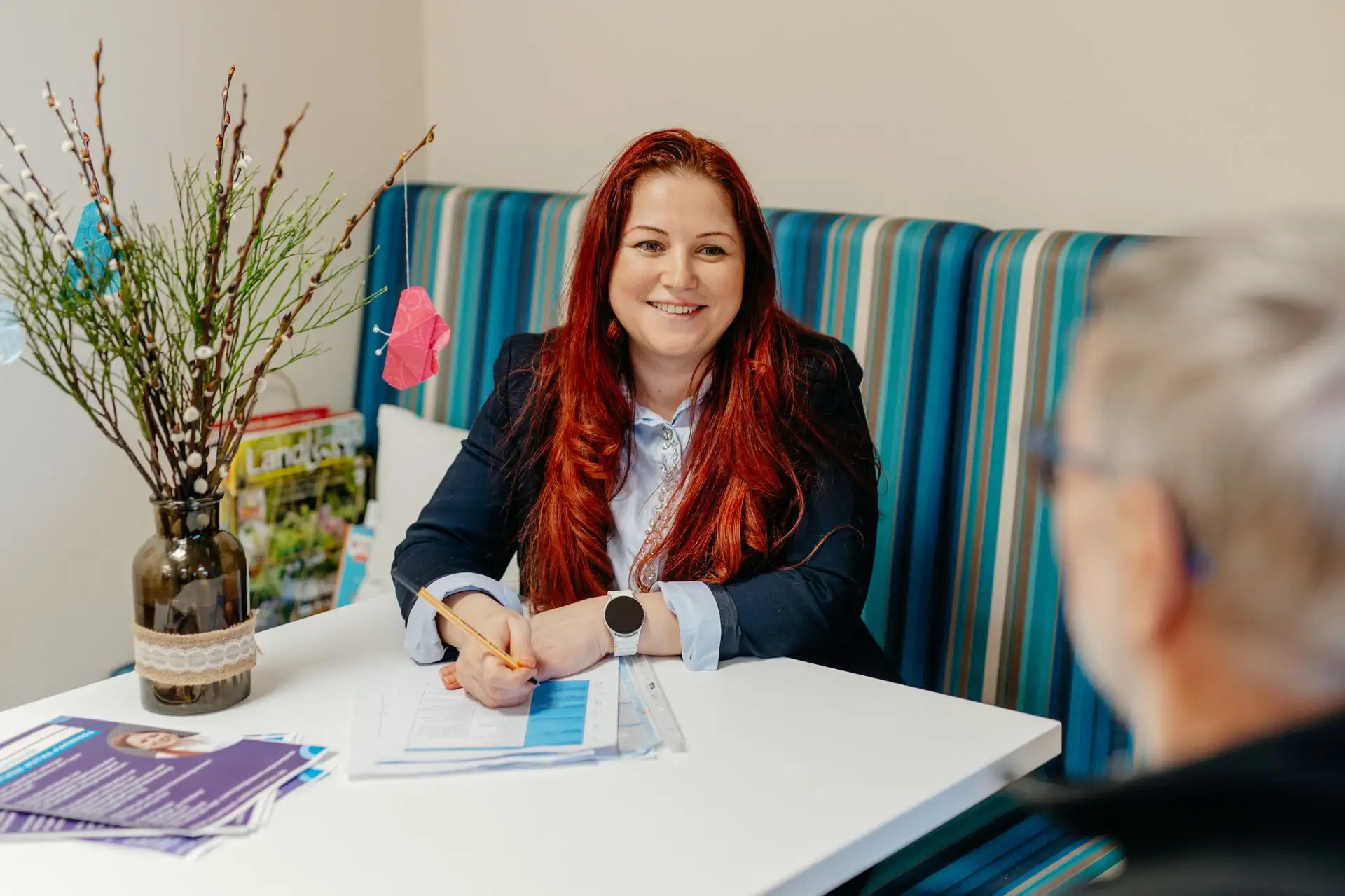 Beraterin mit roten Haaren sitzt an einem Tisch, notiert etwas auf Formularen und lächelt einem älteren Gegenüber zu; im Vordergrund Vase mit Zweigen.
