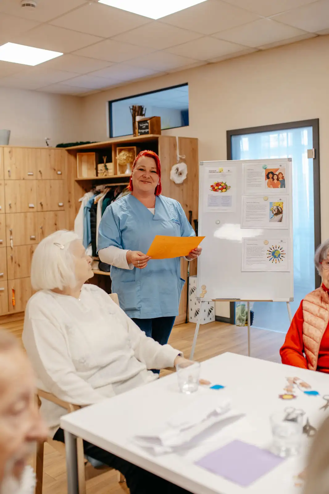 Pflegekraft in blauer Dienstkleidung steht lächelnd mit einem orangefarbenen Blatt vor einer Pinnwand und spricht zu sitzenden Seniorinnen in einem Gemeinschaftsraum.