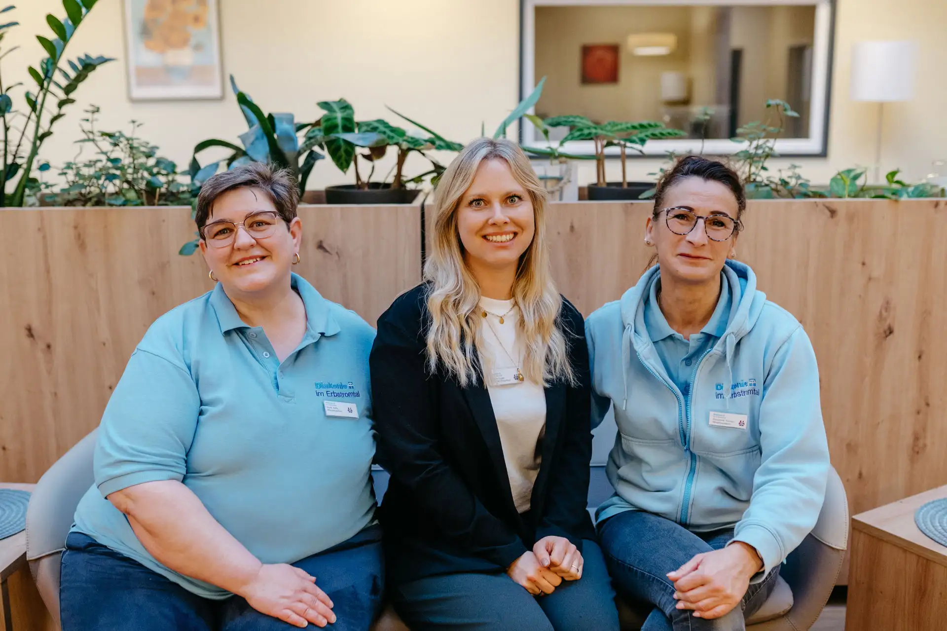 Drei Mitarbeitende sitzen eng nebeneinander vor einer Pflanzwand; zwei tragen hellblaue Diakonie-Polos, die Mitte lächelnde Frau trägt dunkelblazer und Namensschild.