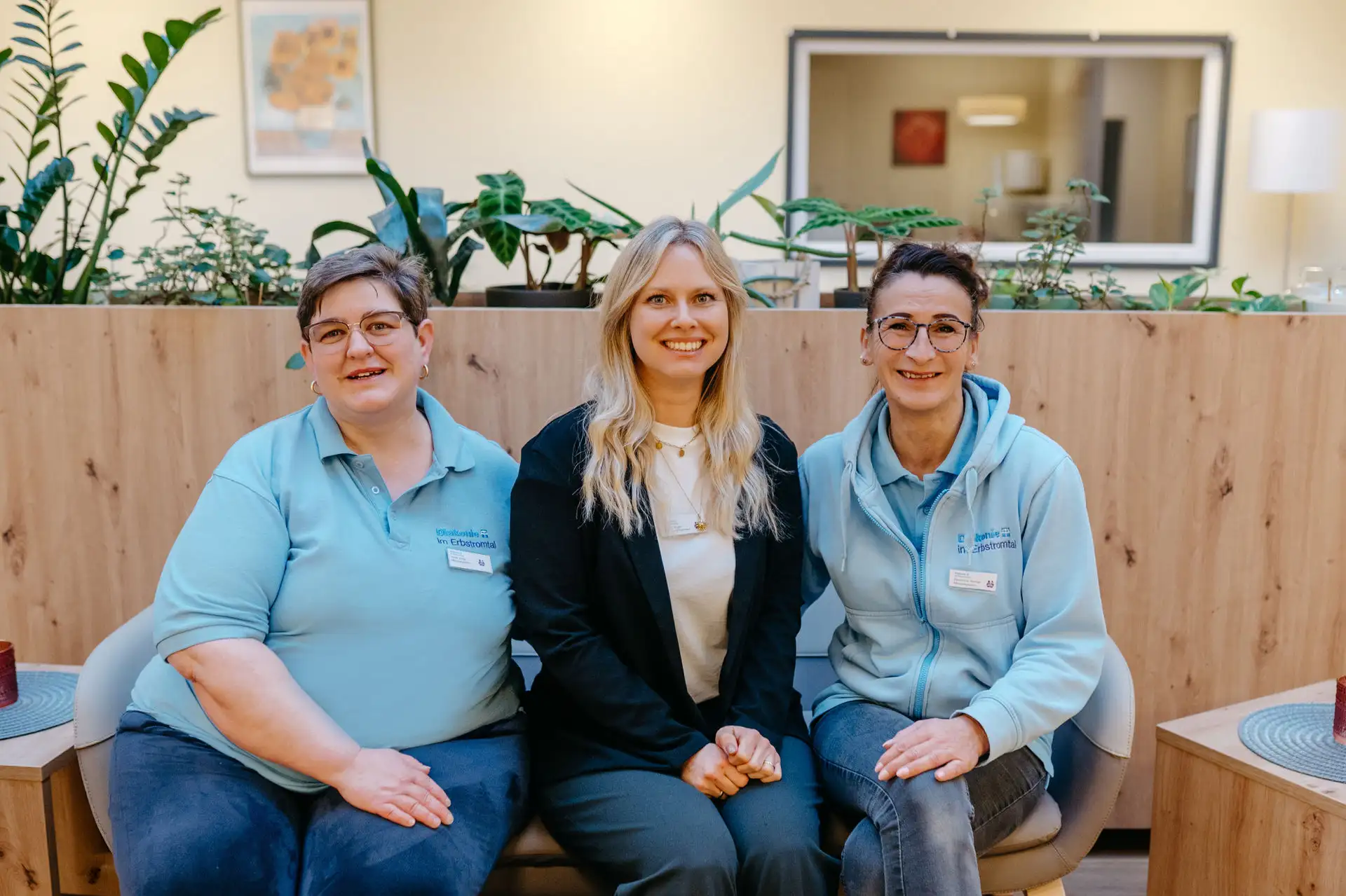 Drei lächelnde Mitarbeitende sitzen nebeneinander in einem hellen Empfangsbereich; zwei tragen Diakonie-Polo/Jacke in Hellblau, die mittlere Person trägt ein schwarzes Sakko, im Hintergrund Pflanzen und Holzverkleidung.