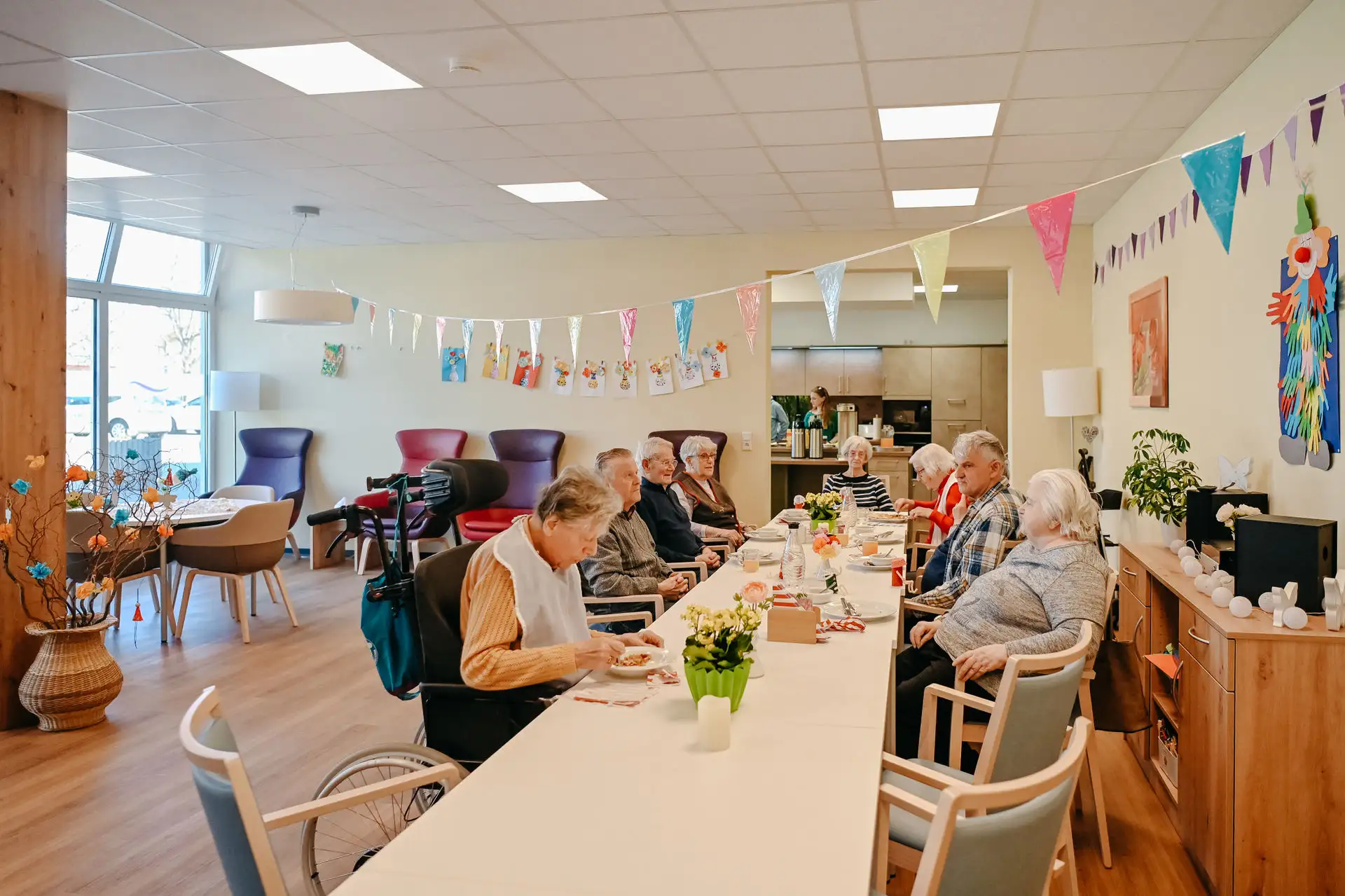 Ältere Menschen sitzen gemeinsam an einem festlich dekorierten Tisch in einem hellen Aufenthaltsraum; Girlanden, Blumen und Getränke deuten auf eine gemütliche Gruppenaktivität hin.
