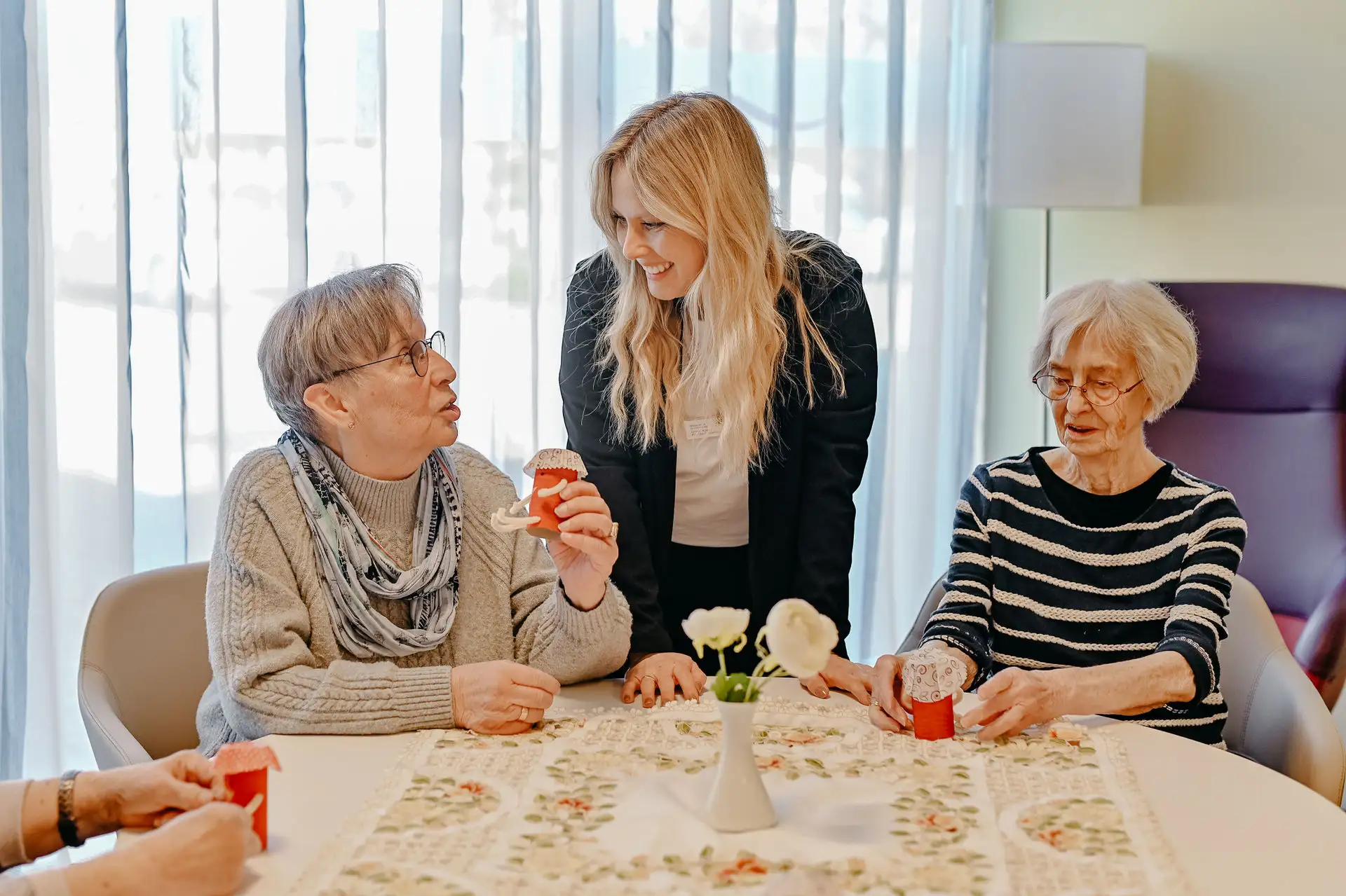 Betreuerin lächelt und spricht mit zwei älteren Frauen, die am Tisch sitzen und basteln; eine hält ein rotes Papiergefäß, auf dem Tisch Blumenvase.