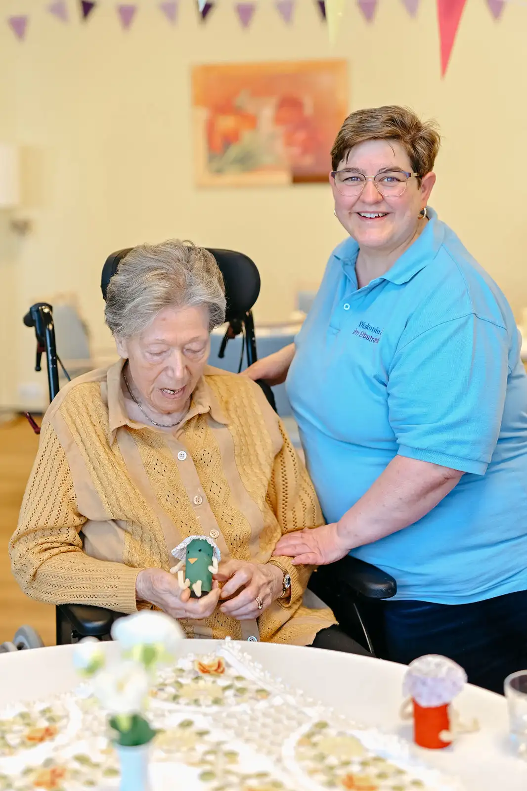 Ältere Frau sitzt im Rollstuhl und hält eine gebastelte Figur, eine Pflegekraft in blauem Polo lächelt und legt unterstützend die Hand auf ihren Arm.