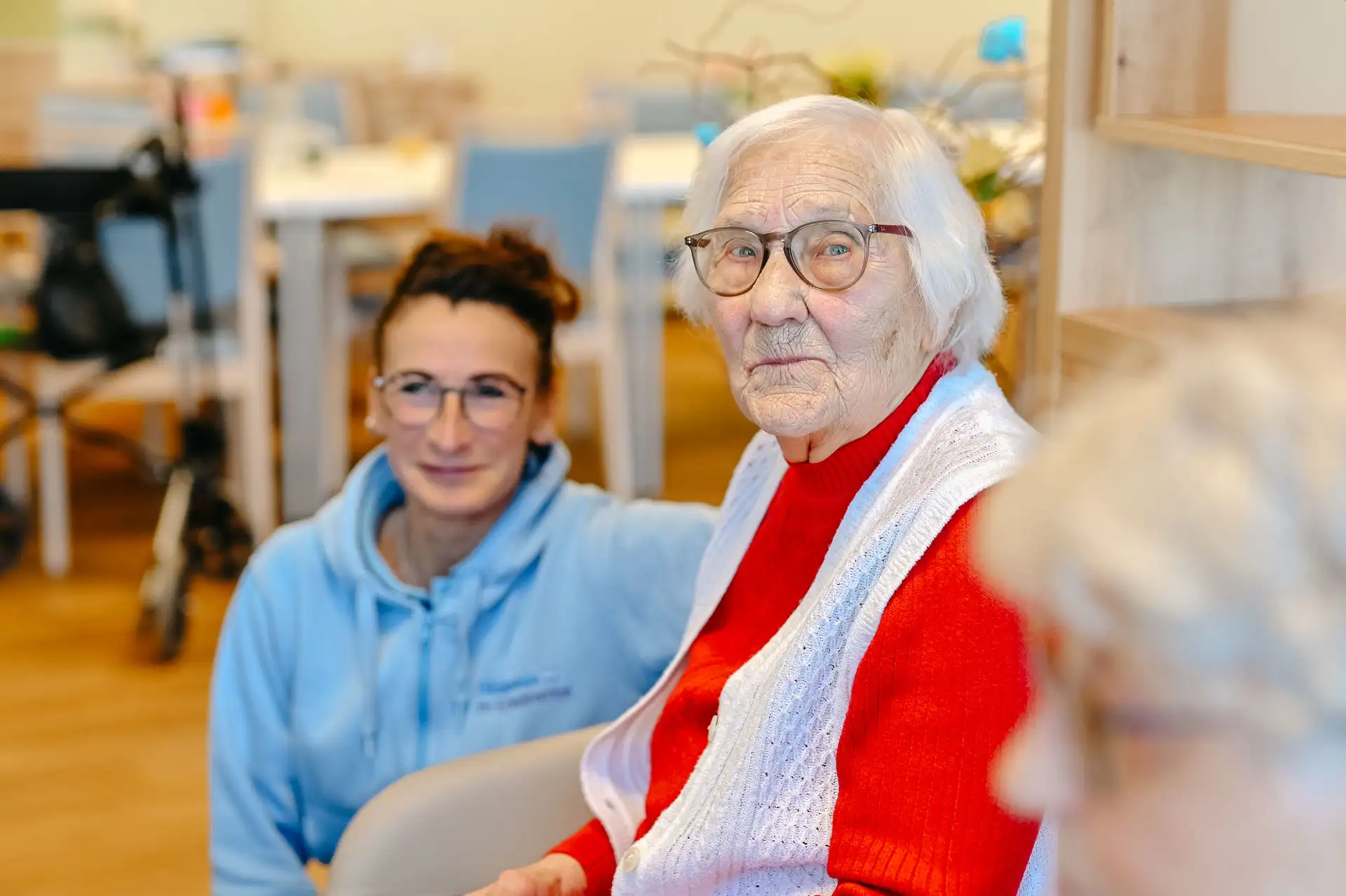 Ältere Frau mit Brille und rotem Pullover sitzt im Wohnbereich, eine Betreuungsperson lächelt im Hintergrund und hält Blickkontakt.