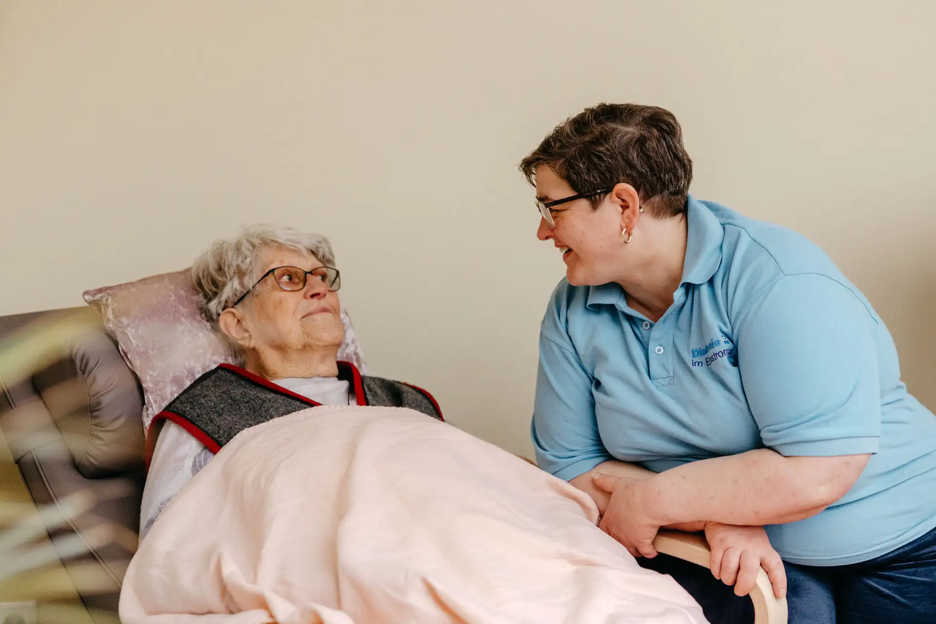 Pflegekraft im hellblauen Poloshirt hält die Hand einer älteren, im Bett liegenden Frau und spricht lächelnd mit ihr; ruhige, fürsorgliche Atmosphäre.