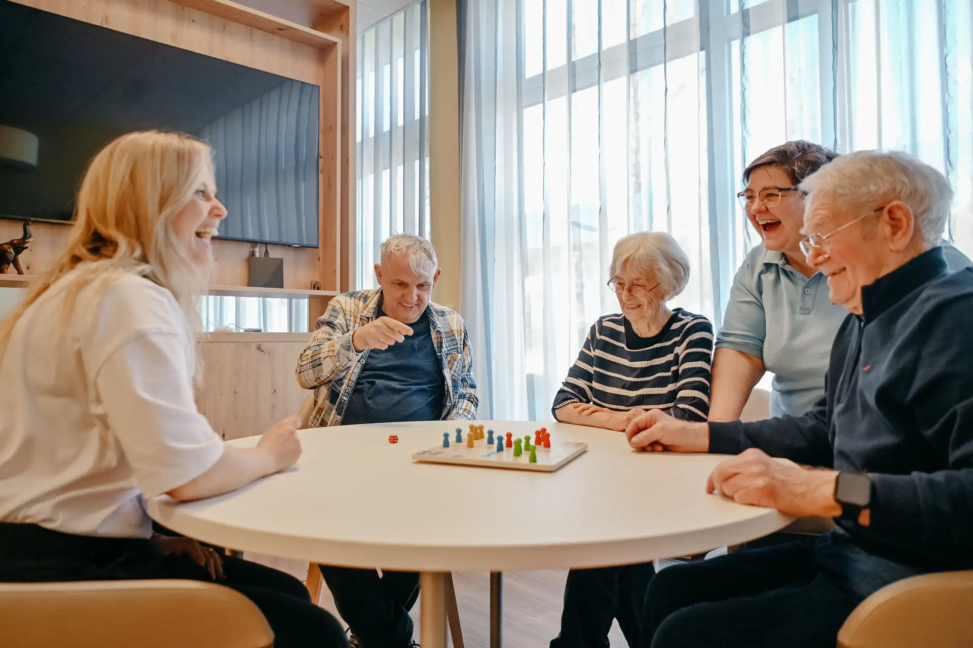 Fünf Menschen unterschiedlichen Alters sitzen an einem runden Tisch und spielen gemeinsam ein Brettspiel; alle lachen und wirken konzentriert und miteinander verbunden.