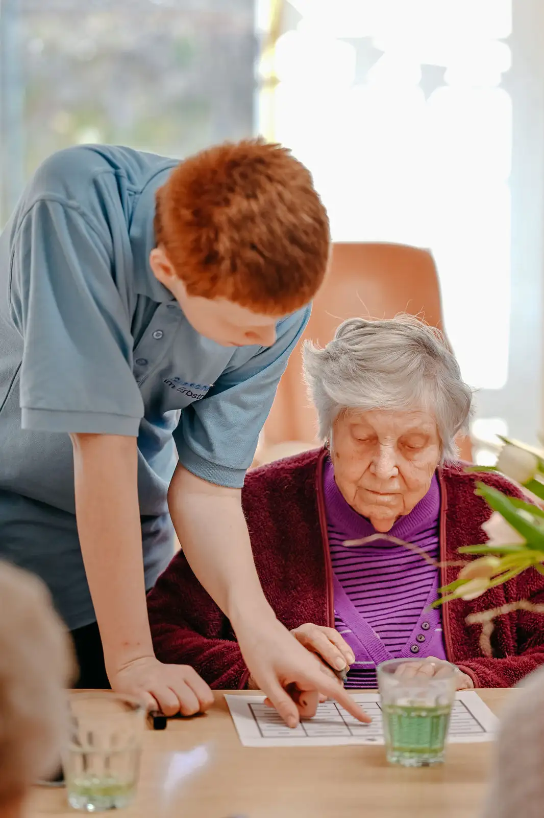 Pflegekraft beugt sich über eine ältere Frau und zeigt ihr auf einem Blatt am Tisch eine Stelle, die Frau blickt konzentriert auf das Papier.