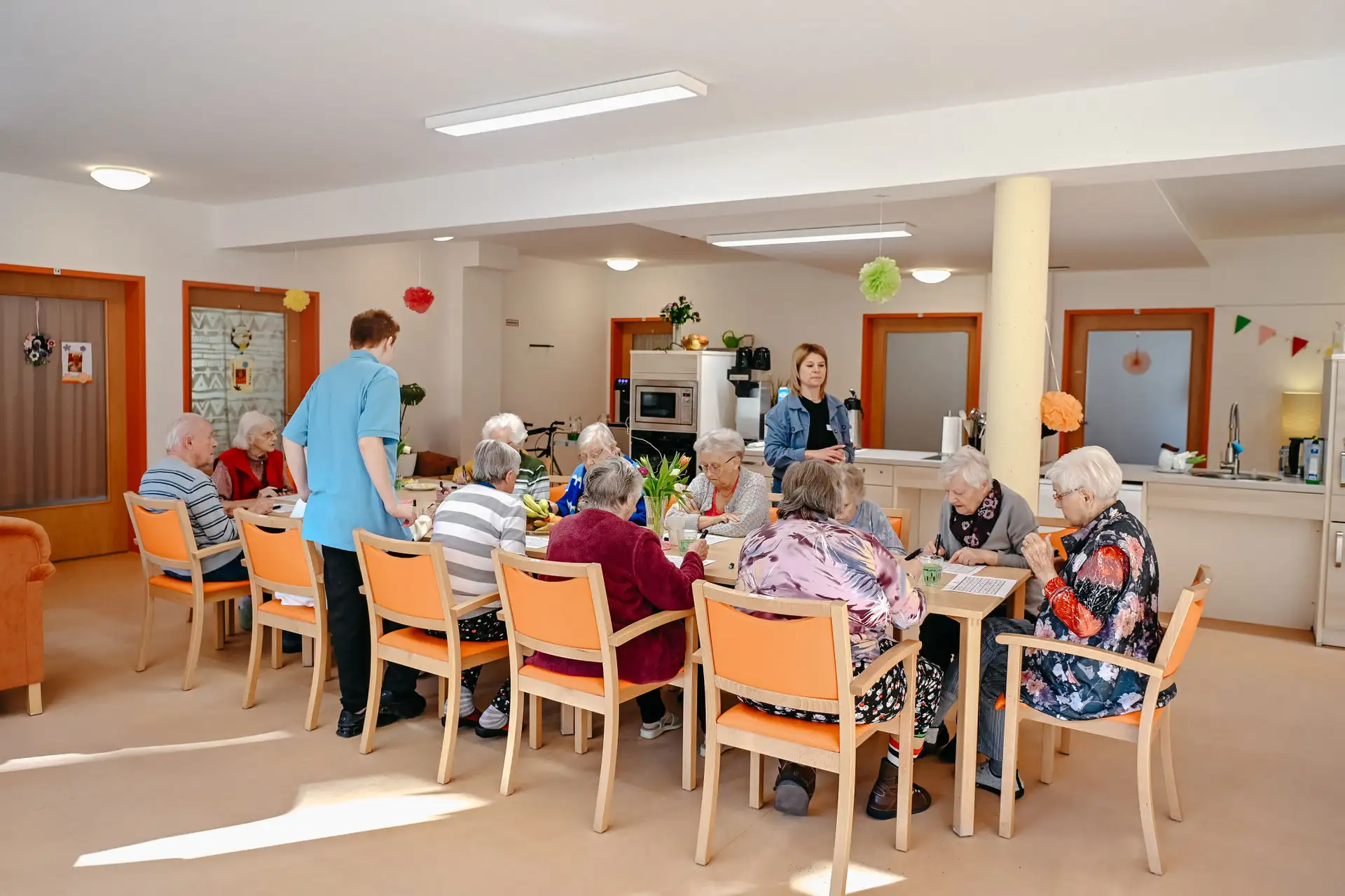 Ältere Menschen sitzen gemeinsam an einem großen Tisch und bearbeiten Rätselblätter, während zwei Betreuungspersonen unterstützen; helle, freundlich dekorierte Gemeinschaftsküche im Hintergrund.