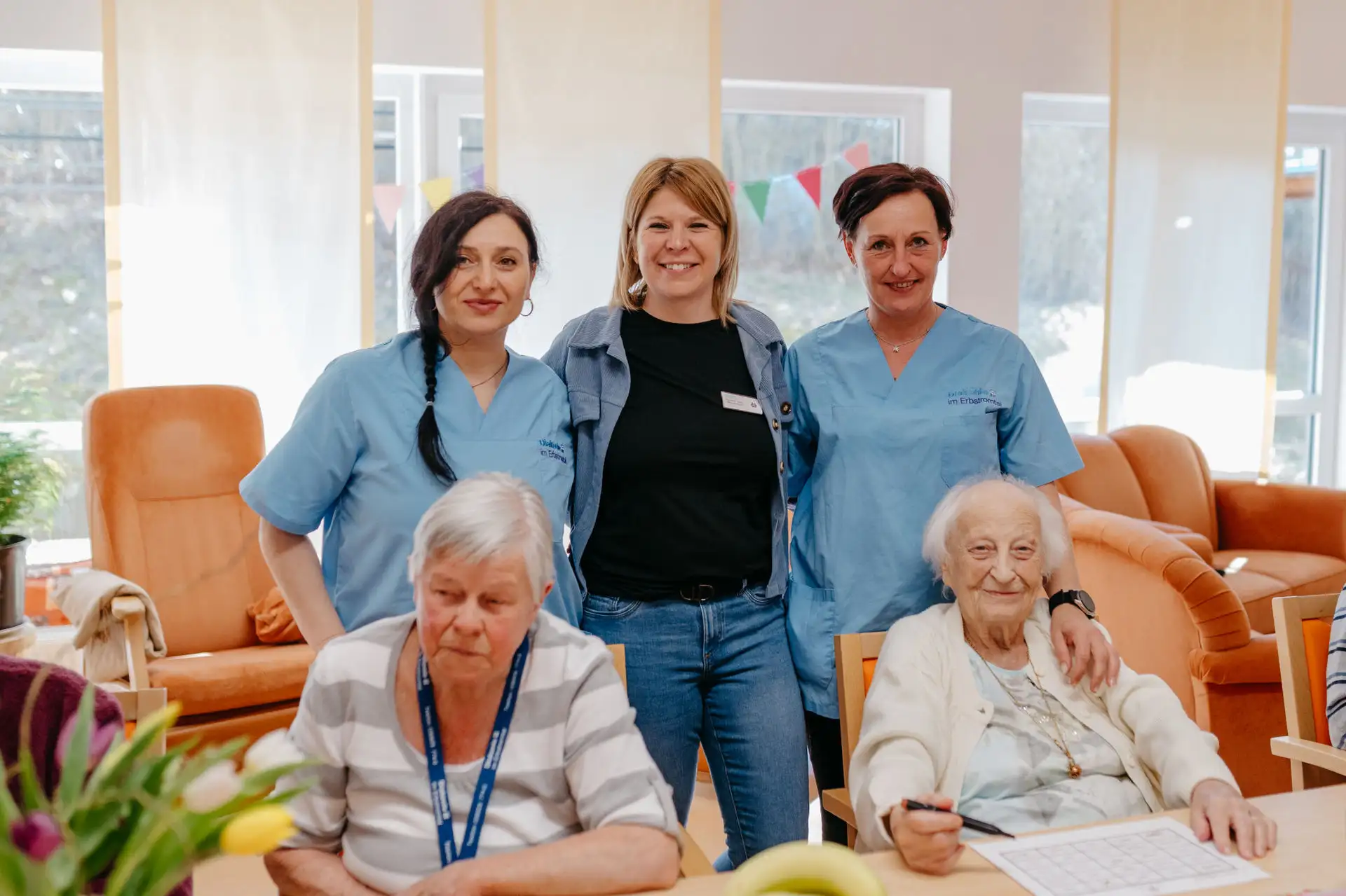 Drei Mitarbeitende stehen lächelnd hinter zwei sitzenden älteren Personen in einem hellen Gemeinschaftsraum; die Ältere rechts hält einen Stift über einem Rätselblatt.