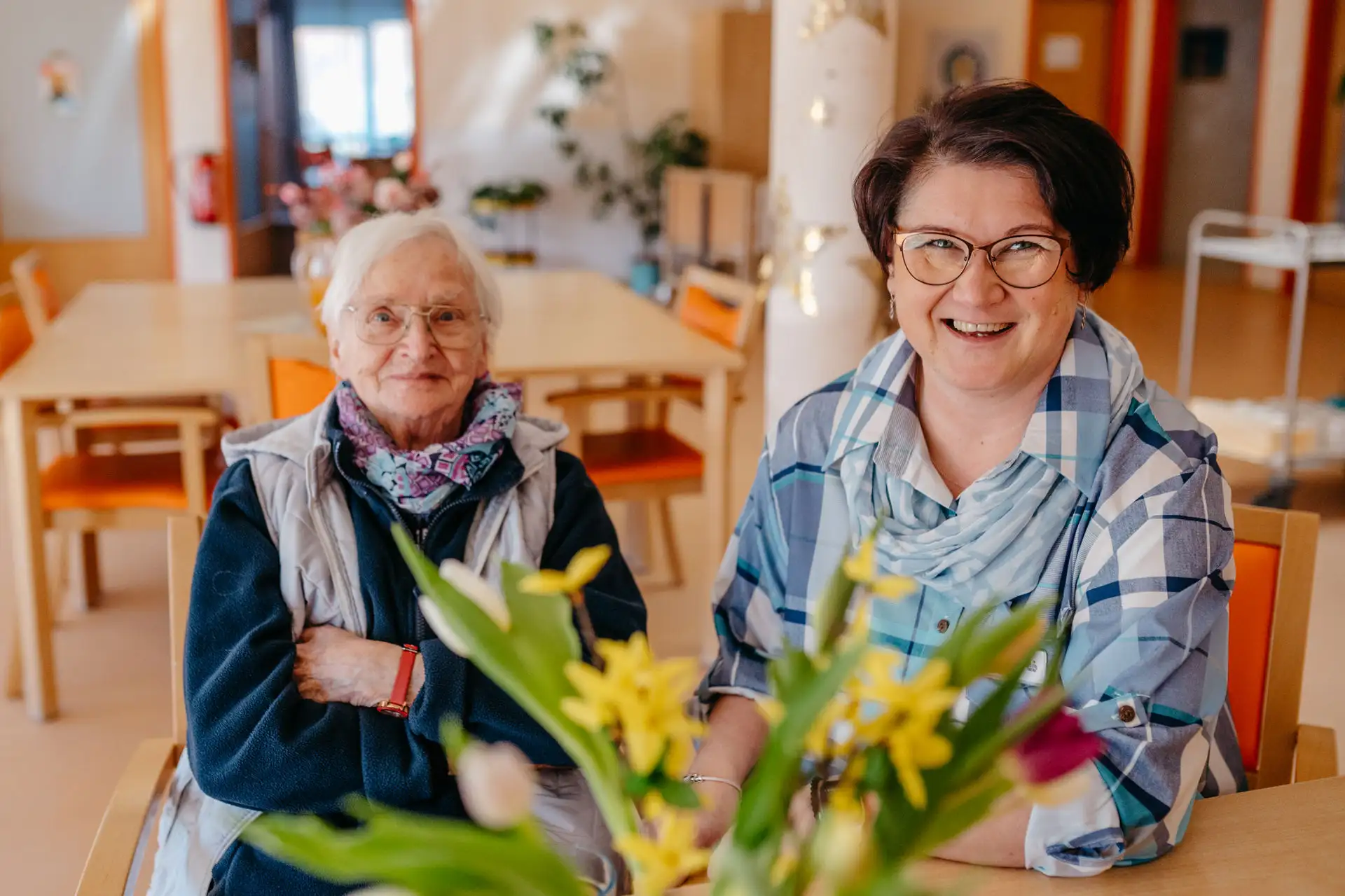 Ältere Frau und Pflegende sitzen nebeneinander an einem Tisch; die Pflegende lächelt offen, im Vordergrund Blumen, im Hintergrund ein Gemeinschaftsraum.
