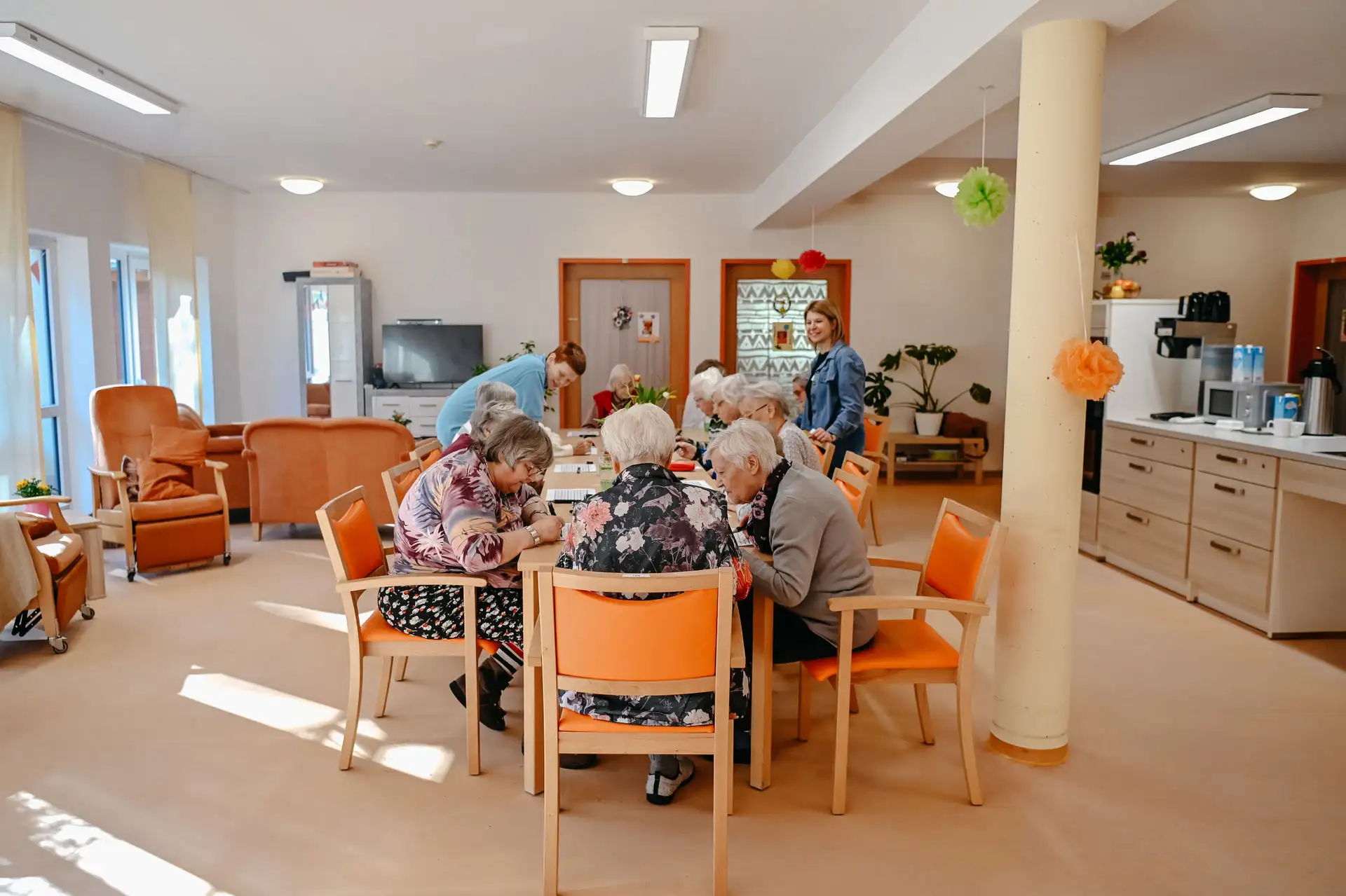 Ältere Menschen sitzen gemeinsam an einem Tisch und arbeiten konzentriert an Papierbögen, während zwei Betreuende im hellen, freundlichen Gemeinschaftsraum Unterstützung anbieten.