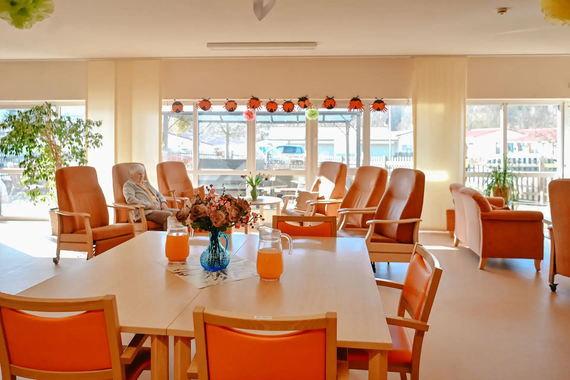 Großer Gemeinschaftsraum mit Reihen von orangefarbenen Sitzstühlen, Tisch mit Blumen und Karaffen, eine ältere Person sitzt entspannt in einem der Sessel.