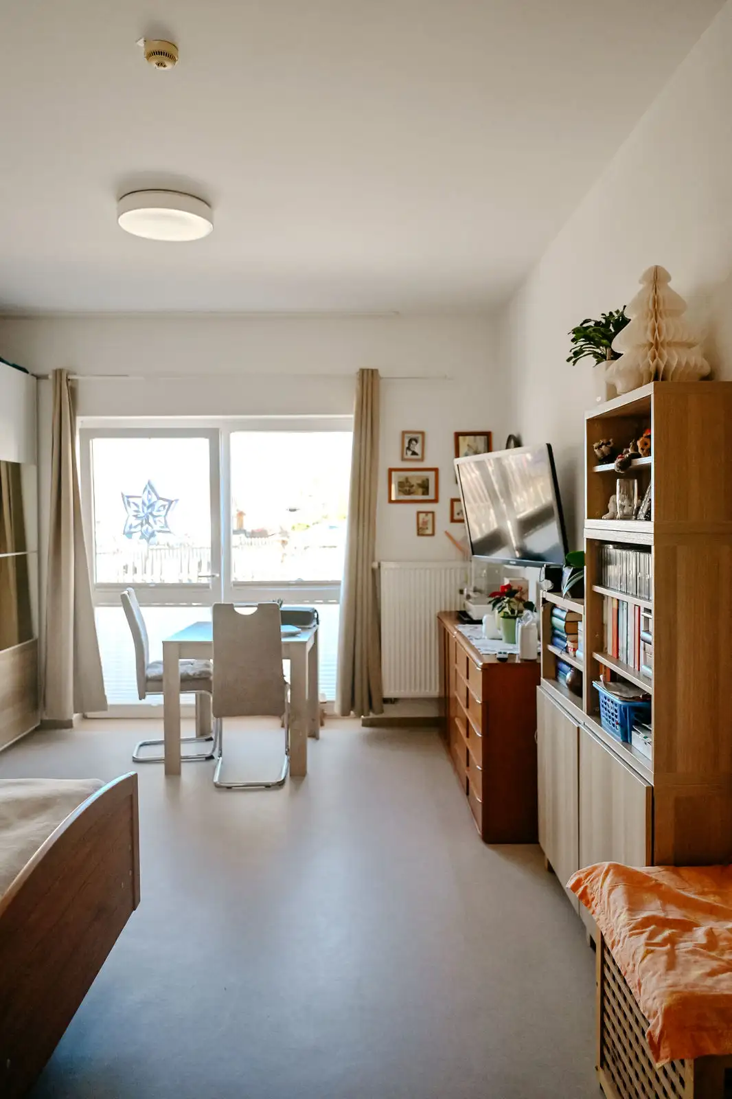 Helles Wohn- und Esszimmer mit Tisch und zwei Stühlen vor großen Fenstern, rechts Regal mit Büchern, Fernseher auf Kommode und dekorativen Pflanzen; ruhige, aufgeräumte Atmosphäre.