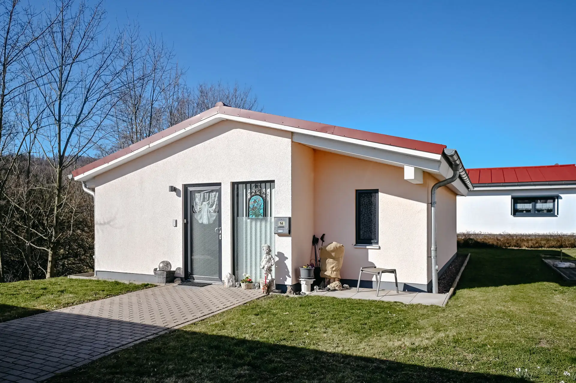 Eingangsbereich eines eingeschossigen Hauses mit rotem Dach; dekorierte Haustür, Briefkasten mit Hausnummer und Gartenstatuen auf dem gepflasterten Weg bei klarem Himmel.