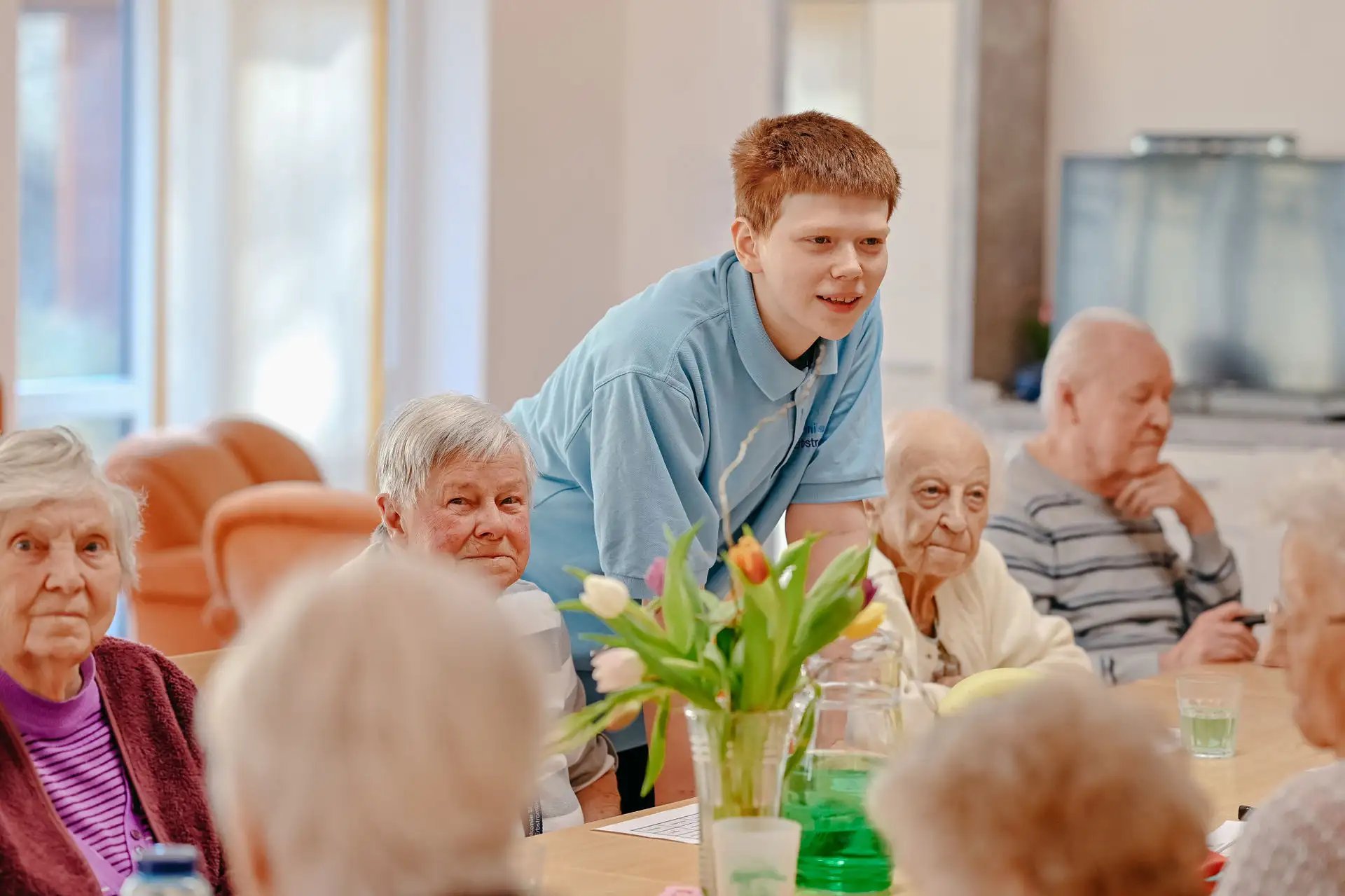 Junge Betreuungsperson in blauem Poloshirt beugt sich lächelnd zu einer Runde älterer Menschen an einem Tisch mit Blumenvasen, offenbar in einem Gemeinschaftsraum einer Pflegeeinrichtung.