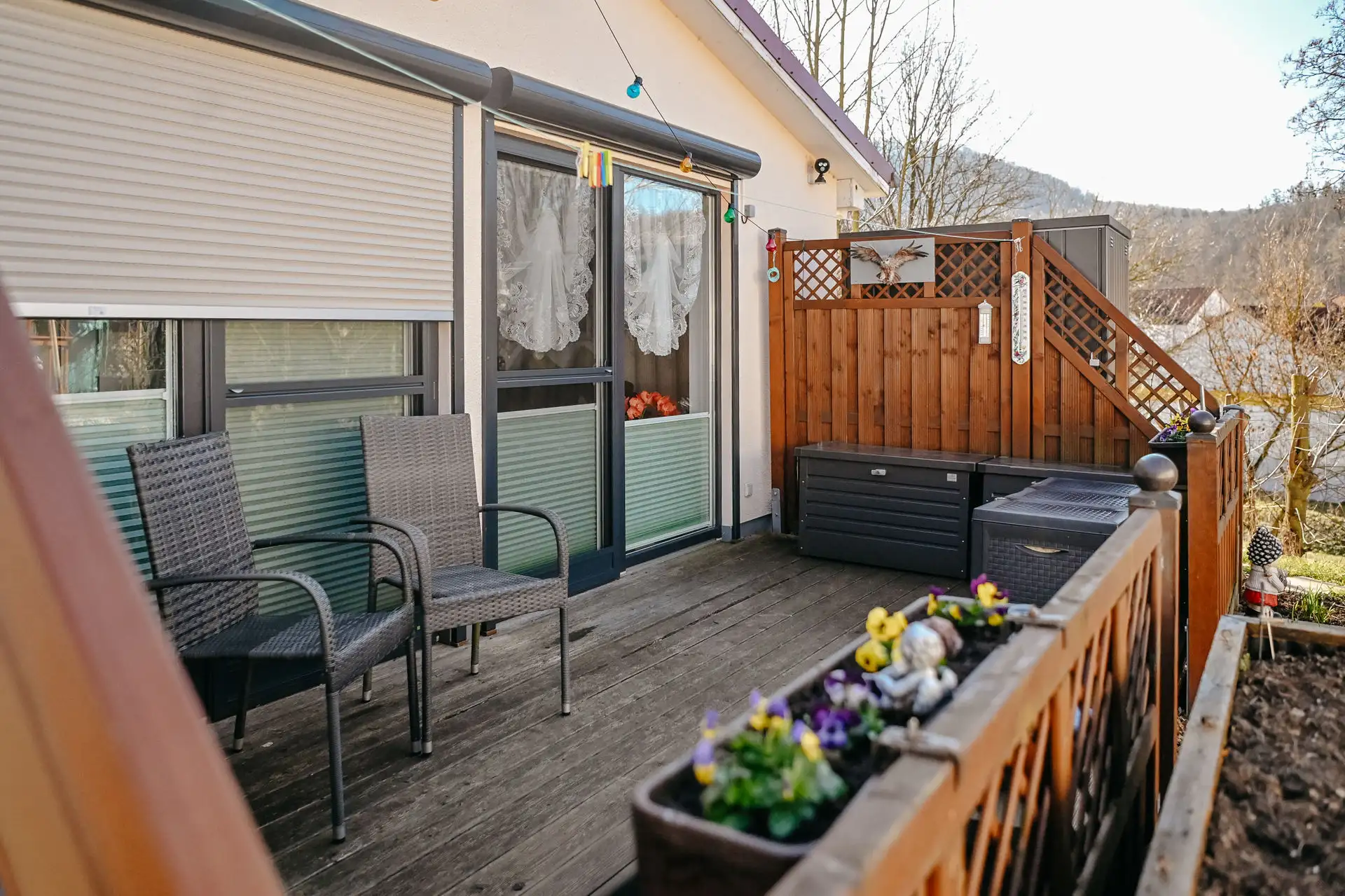 Balkon mit zwei Geflechtstühlen vor einer Balkontür mit Spitzenvorhängen, Blumenkästen am Geländer und Holzsichtschutz mit Dekofigur.