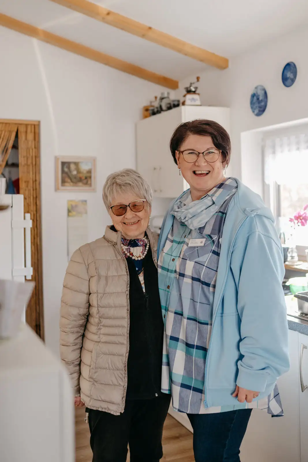 Zwei lächelnde Frauen in einer hellen Küche; die ältere Frau trägt eine gesteppte Jacke und Sonnenbrille, die Begleiterin trägt kariertes Hemd, Namensschild und hellblaue Jacke.
