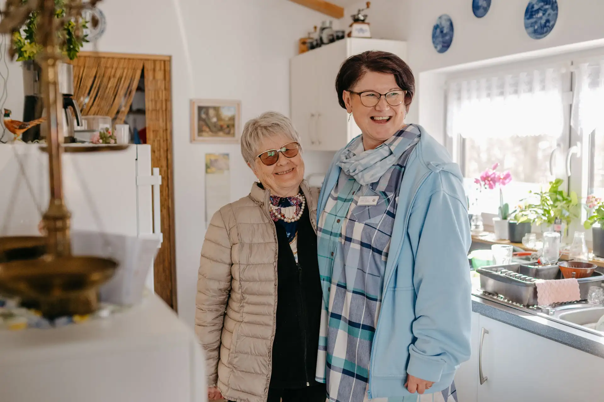 Zwei lächelnde Frauen stehen eng beieinander in einer hellen Küche; die ältere trägt eine helle Steppjacke und Sonnenbrille, die andere eine Brille, blaues Karotuch und Namensschild.