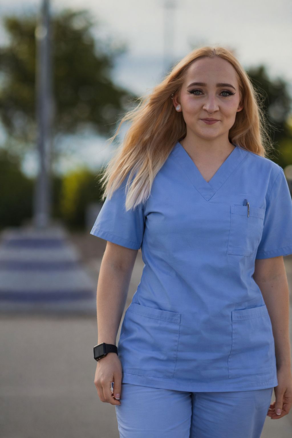 Junge Pflegefachkraft in blauen Kitteln geht nach vorn, lächelt leicht und wirkt freundlich; Haare vom Wind bewegt, Stift in Brusttasche sichtbar.