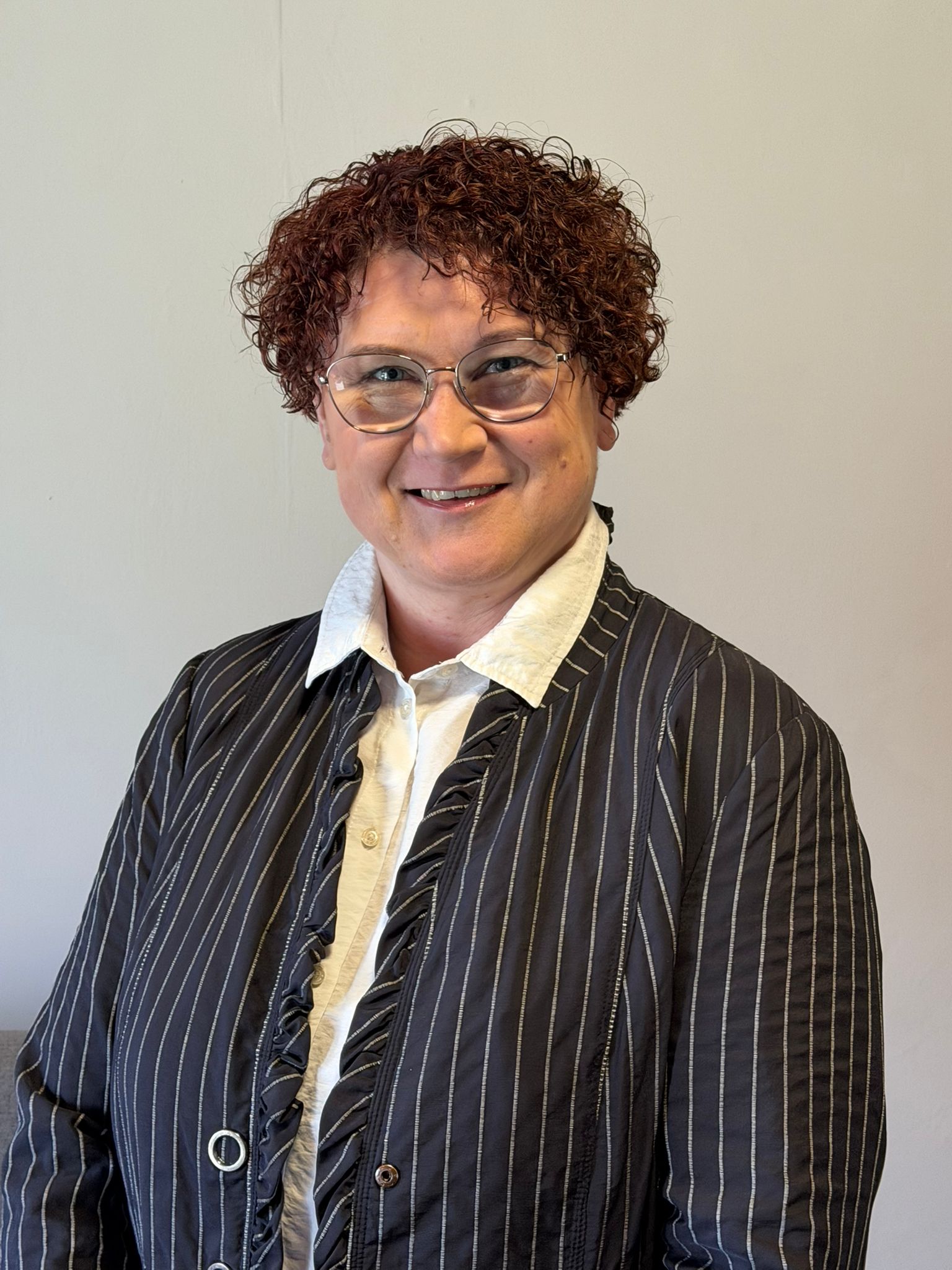 Frau mit kurzen, lockigen roten Haaren und Brille lächelt in die Kamera; sie trägt eine weiße Bluse und eine dunkle, gestreifte Jacke vor neutralem Hintergrund.