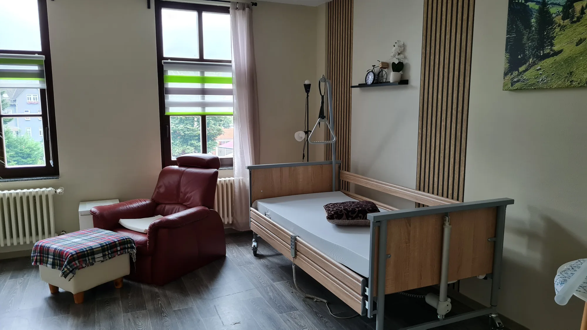Bewohnerzimmer im Haus „Am Thalberg“ mit rotem Sessel, gepolstertem Fußhocker und Pflegebett vor zwei hohen Fenstern mit Blick ins Grüne.